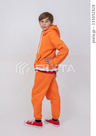 A teenage boy in an orange tracksuit poses on a gray background. 135052828