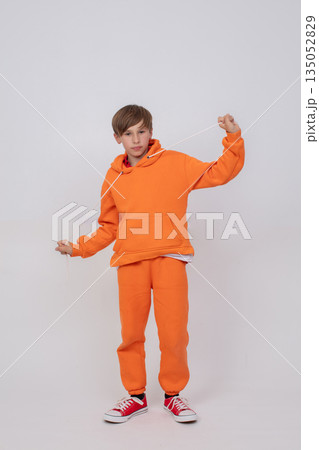 A boy in an orange sweatshirt and orange pants pulls the strings of his hood and looks at the camera. The boy wears red sneakers. A boy in an orange sweatshirt and orange pants pulls the strings of his hood and looks at the camera. The boy wears red sneakers. 135052829
