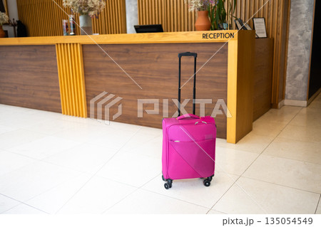 Pink suitcase standing in hotel lobby interior. Travel readiness, movement pause, and modern tourism lifestyle symbolizing transition, journey anticipation, and temporary accommodation experience 135054549