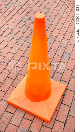 Bright orange plastic traffic cone standing on a red brick sidewalk in an urban area outdoors 135054835