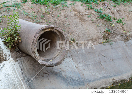 The end of old concrete pipe of large diameter protrudes from the ground above a ditch with a flowing small river with reinforced concreted banks. Road underground communications, symbol of road end 135054904