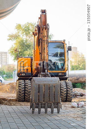 Vertical photo of wheeled excavator at construction site, bucket lowered onto paving slab, front view. Specialized equipment is used for excavation work, laying, replacing pipes, underground utilities Vertical photo of wheeled excavator at construction site, bucket lowered onto paving slab, front view. Specialized equipment is used for excavation work, laying, replacing pipes, underground utilities 135054907