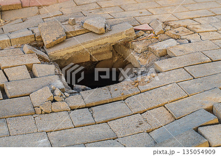 A photograph of a broken sidewalk with fallen paving stones due to a void in the ground. Possible causes include soil collapse and settlement, groundwater erosion, or a burst underground pipeline 135054909
