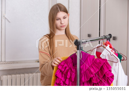 Young girl selecting colorful outfits while organizing clothing rack in a well-lit room during daylight hours Young girl selecting colorful outfits while organizing clothing rack in a well-lit room during daylight hours 135055031