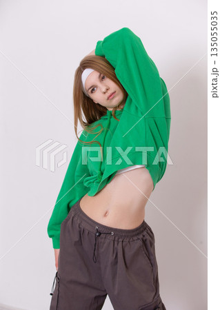 Young woman stretches in a relaxed pose wearing a green hoodie and dark pants against a white background 135055035