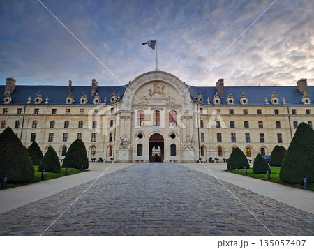 The intricate facade of Les Invalides in Paris. Detailed stone sculptures and the French flag waving above the entrance against a cloudy twilight sky 135057407