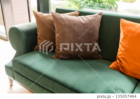 Corner of green sofa with soft pillows near window. Quiet comfort, domestic warmth, and everyday relaxation created through textile texture and natural interior light. Corner of green sofa with soft pillows near window. Quiet comfort, domestic warmth, and everyday relaxation created through textile texture and natural interior light. 135057464