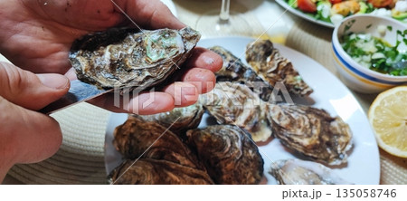 A close-up of hands opening fresh oysters with a knife over a plate. Concepts of fine dining, seafood delicacies, restaurant dining, home cooking, nutrition, and authentic gastronomic experiences. A close-up of hands opening fresh oysters with a knife over a plate. Concepts of fine dining, seafood delicacies, restaurant dining, home cooking, nutrition, and authentic gastronomic experiences. 135058746