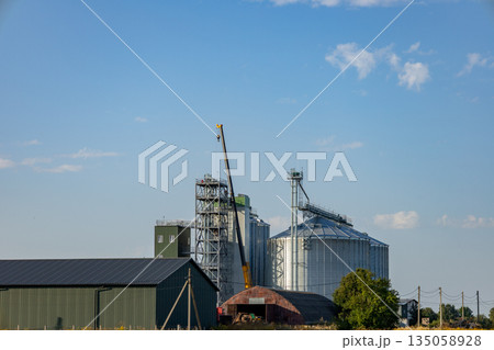 Cranes hover above large silos in a bustling granary, showcasing the harmony of agricultural storage and industrial activity in a serene landscape Cranes hover above large silos in a bustling granary, showcasing the harmony of agricultural storage and industrial activity in a serene landscape 135058928
