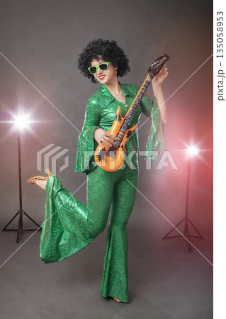 Fun eighties style. Girl comic pop star posing in black wig with toy guitar on gray background. Fun eighties style. Girl comic pop star posing in black wig with toy guitar on gray background. 135058953