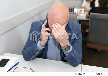 A tired businessman is talking on his smartphone and he covered his face with his hands. 135058957