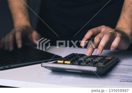 Man's hands are pressing buttons on a calculator, calculating expenses on an invoice and saving the data onto a laptop. Man's hands are pressing buttons on a calculator, calculating expenses on an invoice and saving the data onto a laptop. 135058987