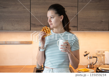 Woman eating waffle and holding cup with drink in home kitchen. Everyday breakfast moment and relaxed food routine in real life interior Woman eating waffle and holding cup with drink in home kitchen. Everyday breakfast moment and relaxed food routine in real life interior 135059234