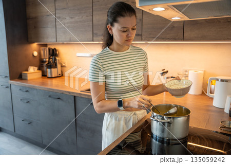 A woman pours soup from a pot into a bowl while cooking in a home kitchen. Real life interior scene showing everyday meal preparation, domestic routine and homemade food. A woman pours soup from a pot into a bowl while cooking in a home kitchen. Real life interior scene showing everyday meal preparation, domestic routine and homemade food. 135059242