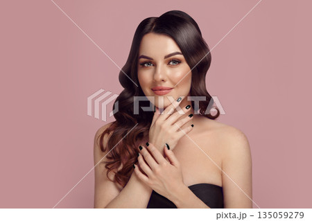 Brunette caucasian woman with a beautiful nude makeup and wavy hair, having a black manicure putting her hands to the face posing over pink isolated background. Brunette caucasian woman with a beautiful nude makeup and wavy hair, having a black manicure putting her hands to the face posing over pink isolated background. 135059279