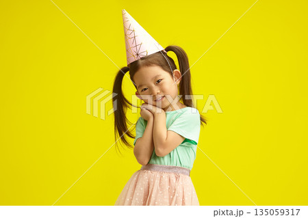 Asian little girl in party hat smiling coyly, cute placed palms of hands in front of herself face, looks like as if begging for a desired gift or makes a wish on herself birthday, stands over yellow 135059317
