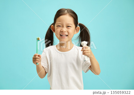 Beautiful asian ethnicity kid girl holding toothbrush and toothpaste tube, wearing in white t shirt, having expression joyful, preparing cleans her teeth, standing over blue isolated background. Beautiful asian ethnicity kid girl holding toothbrush and toothpaste tube, wearing in white t shirt, having expression joyful, preparing cleans her teeth, standing over blue isolated background. 135059322