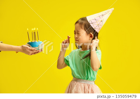 Chinese Little Kid Girl Wishing on Birthday Candles Stands Against Yellow Isolated Background, Closing her Eyes and Crossing Fingers, Wearing in Party Hat, Expressing Hope for Wish Fulfillment  135059479