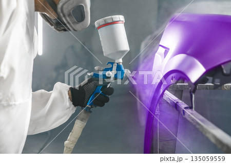 Car paint worker spraying purple paint to car body element using spray gun in vehicle workshop chamber. Complex restoration of automobile body. Car paint worker spraying purple paint to car body element using spray gun in vehicle workshop chamber. Complex restoration of automobile body. 135059559