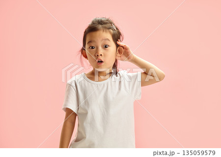 amazed little girl has curiosity putting hand to ear and overhear something expresses surprise, can't believe what she heard standing on pink isolated background amazed little girl has curiosity putting hand to ear and overhear something expresses surprise, can't believe what she heard standing on pink isolated background 135059579