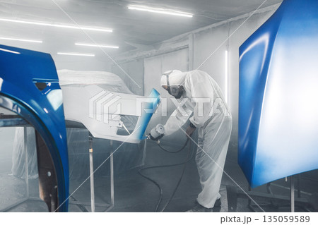 Car paint worker spraying blue paint to car body element using spray gun in vehicle workshop chamber. Complex restoration of automobile body.  135059589