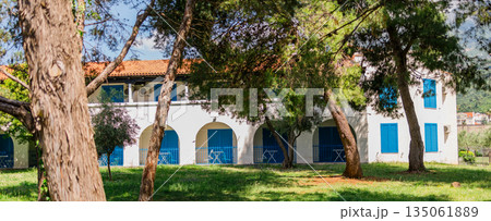 White building with blue shutters and arches surrounded by garden. Mediterranean architecture, serene vacation atmosphere, and hotel patio concept. 135061889