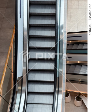 Escalators inside a contemporary shopping center viewed from above. Movement, symmetry, and urban architecture symbolizing daily routine and structure. Escalators inside a contemporary shopping center viewed from above. Movement, symmetry, and urban architecture symbolizing daily routine and structure. 135064262