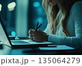 Woman working at laptop in a dimly lit office during late hours with pens and notepad 135064279