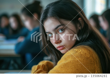 Student in classroom wearing yellow sweater gazes thoughtfully while peers focus on their work 135064281