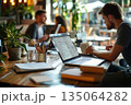 Young man working intently at a laptop in a lively cafe filled with indoor plants and soft ambient lighting 135064282