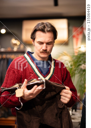 Fashionable man customer purchasing clothing in vintage men's shop, picking trendy velvet pants. Fashionable man customer purchasing clothing in vintage men's shop, picking trendy velvet pants. 135064419