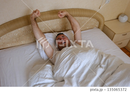 man stretching on bed in cozy bedroom with soft lighting and minimalistic decor featuring books and a small lamp 135065372