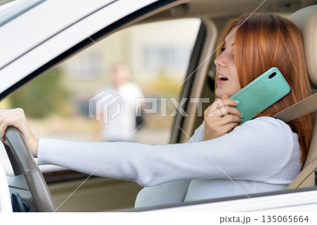 Happy redhead girl talking on her mobile phone behind the wheel driving a car. 135065664