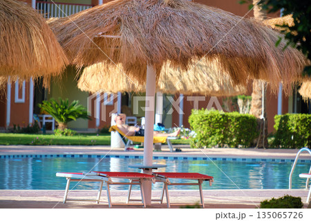 Empty deck chairs under straw shade umbrellas on swimming pool side in tropical resort. Summer vacations and getaway concept Empty deck chairs under straw shade umbrellas on swimming pool side in tropical resort. Summer vacations and getaway concept 135065726