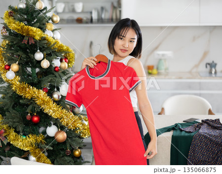 Happy young Asian girl trying to choose a dress standing next to Xmas-tree 135066875