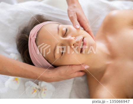 Young female client relaxing in spa salon during massage of face Young female client relaxing in spa salon during massage of face 135066947
