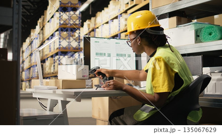 Black female with hi vis vest registering awb tracking info with scanner, oversees express delivery service for cargo orders. Scanning shipment details on labels, monitoring pc software. Camera A. Black female with hi vis vest registering awb tracking info with scanner, oversees express delivery service for cargo orders. Scanning shipment details on labels, monitoring pc software. Camera A. 135067629