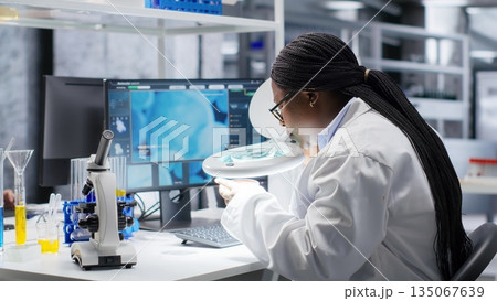 Woman researcher conducts observation of Petri dish under magnifying glass in lab. Analysis of microorganisms and bacterial growth highlights experimental methods, biotechnology. Woman researcher conducts observation of Petri dish under magnifying glass in lab. Analysis of microorganisms and bacterial growth highlights experimental methods, biotechnology. 135067639