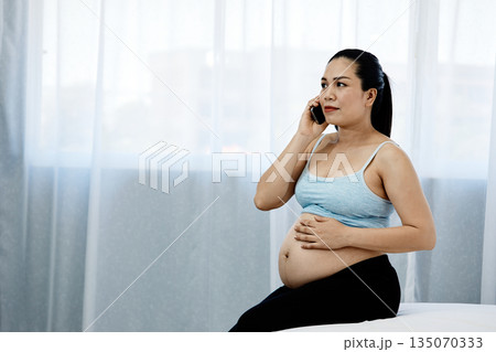 Pregnant Asian woman sitting on bed, holding her belly and talking on the phone with calm expression, representing motherhood, prenatal care, and healthy pregnancy lifestyle at home Pregnant Asian woman sitting on bed, holding her belly and talking on the phone with calm expression, representing motherhood, prenatal care, and healthy pregnancy lifestyle at home 135070333