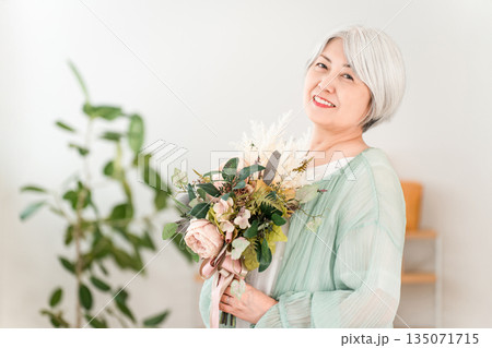 自宅のリビングで花束を持つグレイヘアーのシニア女性 135071715