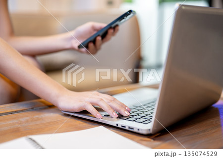 Close up woman hand holding phone aside laptop and notebook on table sitting at sofa in living room Close up woman hand holding phone aside laptop and notebook on table sitting at sofa in living room 135074529