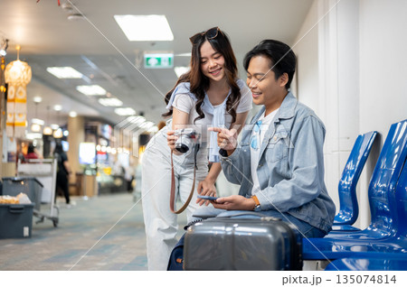 Asian tourist couple go on trip a woman showing camera to man while waiting for departure in airport Asian tourist couple go on trip a woman showing camera to man while waiting for departure in airport 135074814