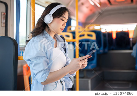 Asian woman tourist wear headphones looking at phone standing leaning against a pole in shuttle bus. Asian woman tourist wear headphones looking at phone standing leaning against a pole in shuttle bus. 135074860