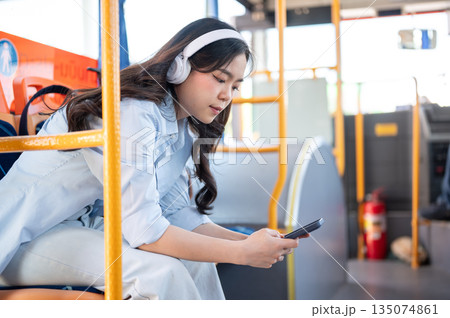 Asian woman tourist wear headphones listening to music looking at phone while sitting in shuttle bus Asian woman tourist wear headphones listening to music looking at phone while sitting in shuttle bus 135074861