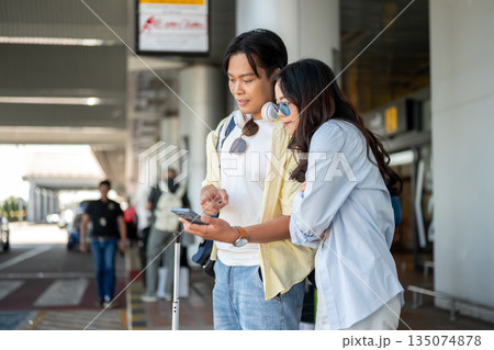 Asian tourist couple go on trip woman looking at man's phone as standing waiting for taxi in airport Asian tourist couple go on trip woman looking at man's phone as standing waiting for taxi in airport 135074878