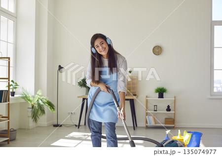Housewife in earphones listening to music vacuuming the floor with vacuum cleaner at home. 135074977