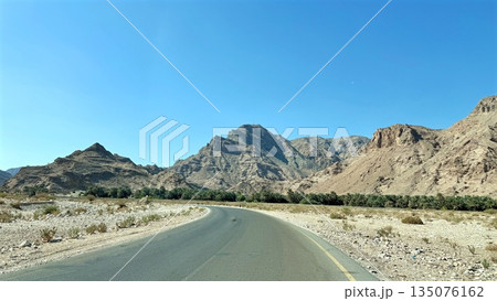 Sunlit Road Through Barren Mountains, Vast Highway In Dry Valley With Distant Summits, An Expansive Deserted Road Traverses Dry Mountain Depression With Panoramic Blue Skies And Minimal Plant Life Sunlit Road Through Barren Mountains, Vast Highway In Dry Valley With Distant Summits, An Expansive Deserted Road Traverses Dry Mountain Depression With Panoramic Blue Skies And Minimal Plant Life 135076162