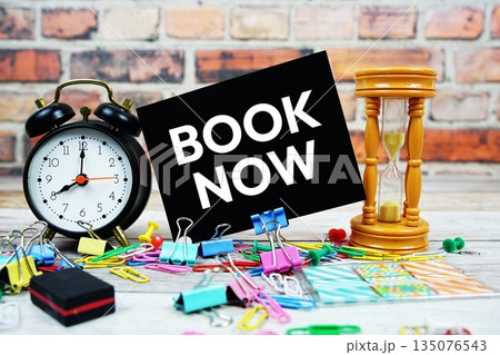 Book Now text on paper card with alarm clock and stationary on wooden background 135076543