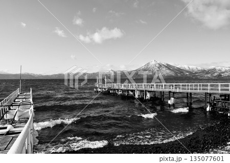 「1月の支笏湖」凍った桟橋に打ち寄せる波　奥に雪をまとった「恵庭岳」白黒 135077601
