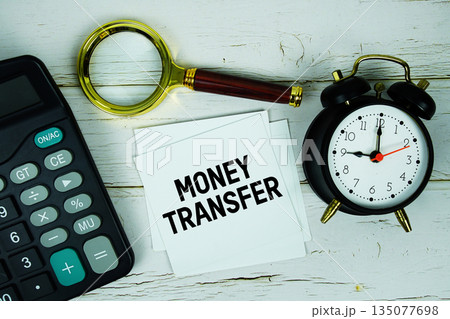 Money Transfer text with calculator, magnifying glass and alarm clock on wooden background 135077698